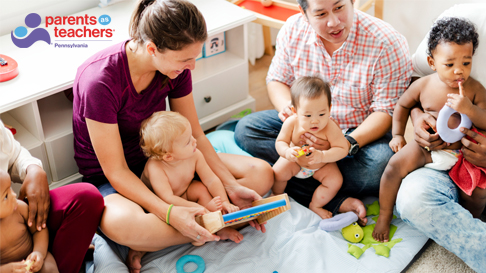 Father and his young child are on the floor surrounded by pillows and balls and a teepee is in the background. He is reading a story to his child, who is looking at him and both are laughing. They are wearing homemade cardboard crowns.